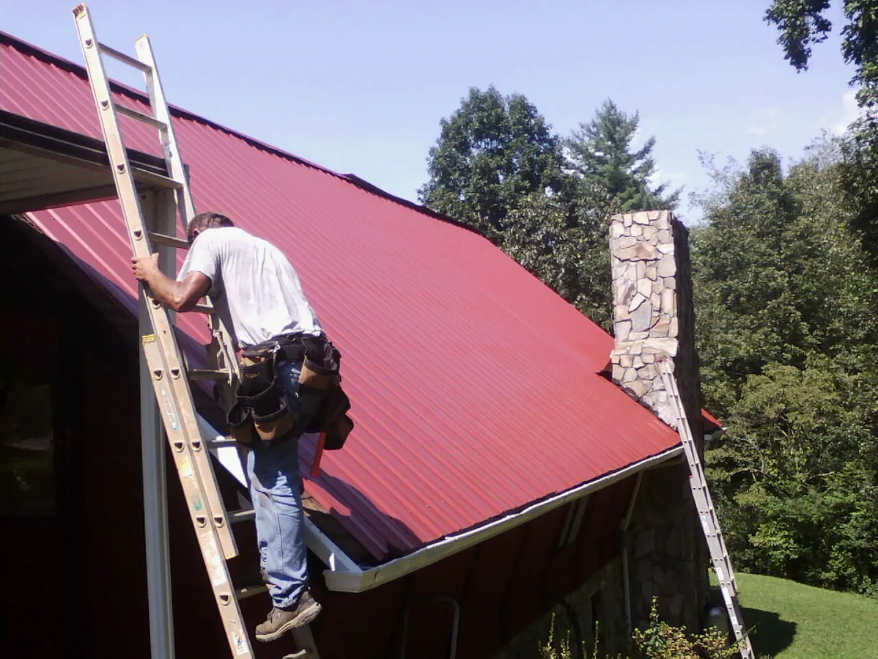 Roofer on a ladder beside a red metal roof and stone chimney — Soffit & Fascia Repair by Roofing Brothers in Lonedell